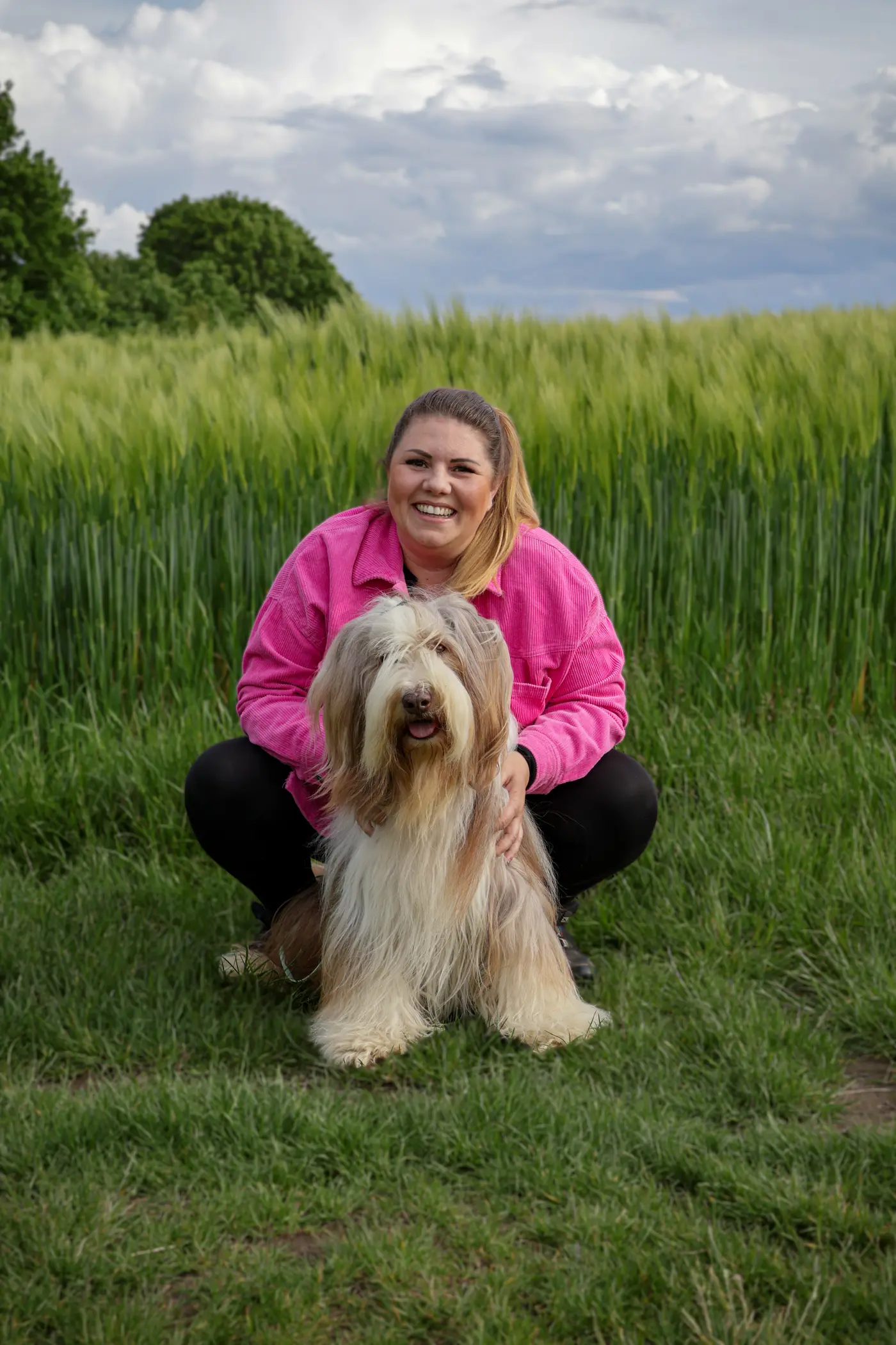 Laura hockt lachend hinter ihrem sitzenden Bearded Collie